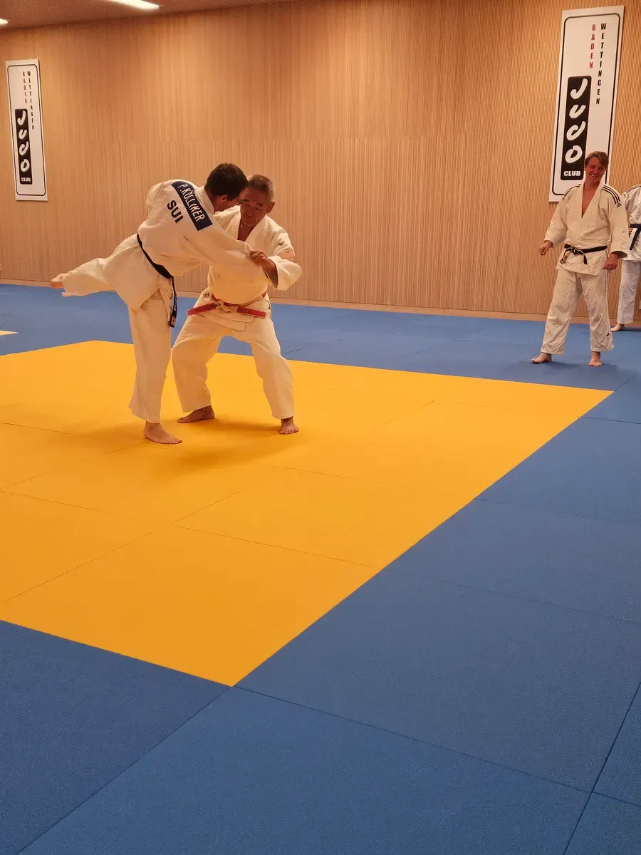 Hiroshi Katanishi zeigt mit einem Partner auf der gelben Wettkampffläche im Dojo Baden-Wettingen, wie Bewegung und Gleichgewicht zusammenspielen